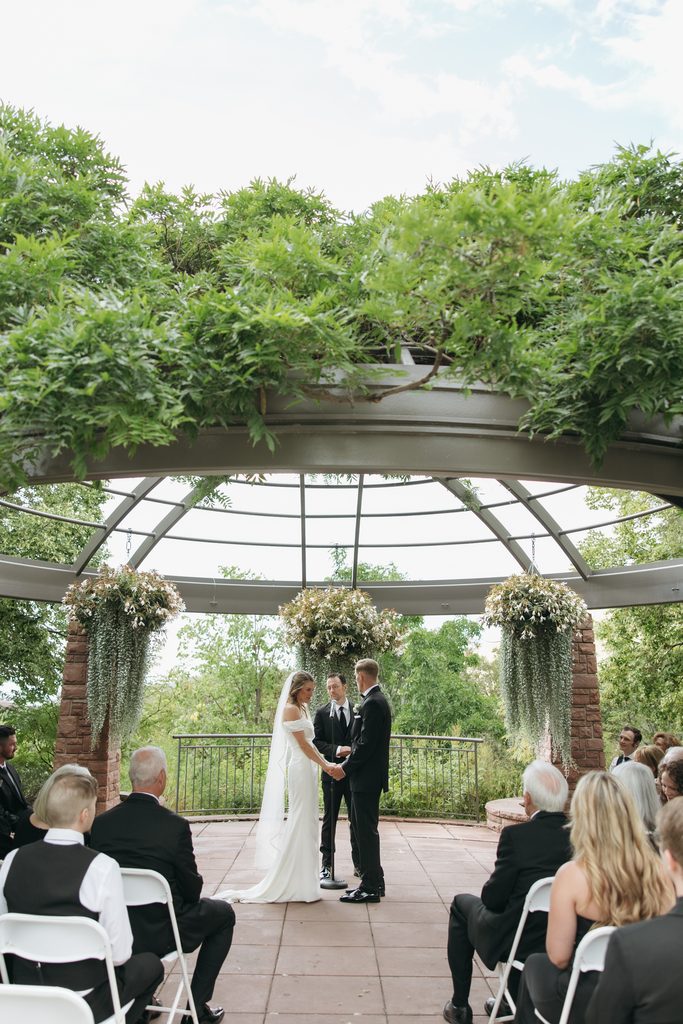 A and M holding hands in front of guests during ceremony at Red Butte Garden, surrounded by greenery.