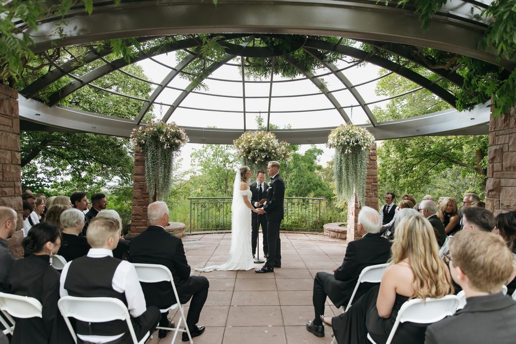 A and M holding hands in front of guests during ceremony at Red Butte Garden, surrounded by greenery.