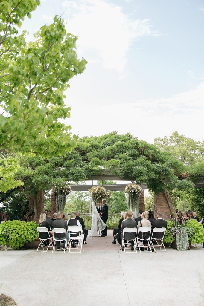 A and M holding hands in front of guests during ceremony at Red Butte Garden, surrounded by greenery.