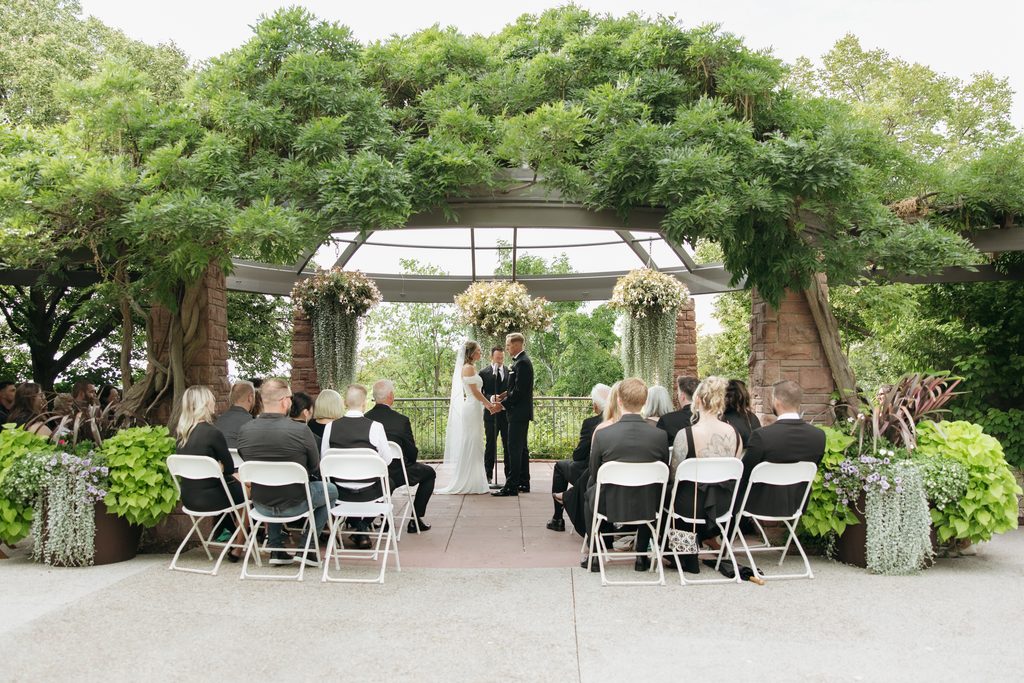 A and M holding hands in front of guests during ceremony at Red Butte Garden, surrounded by greenery.