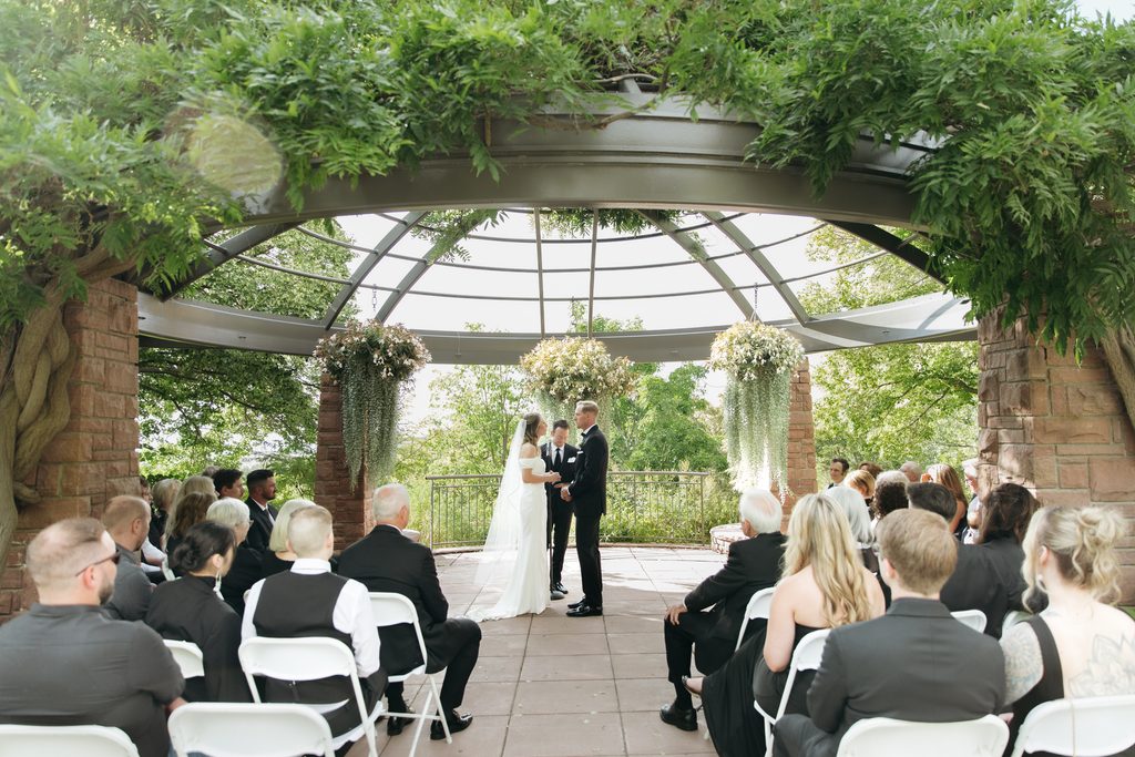 A and M holding hands in front of guests during ceremony at Red Butte Garden, surrounded by greenery.